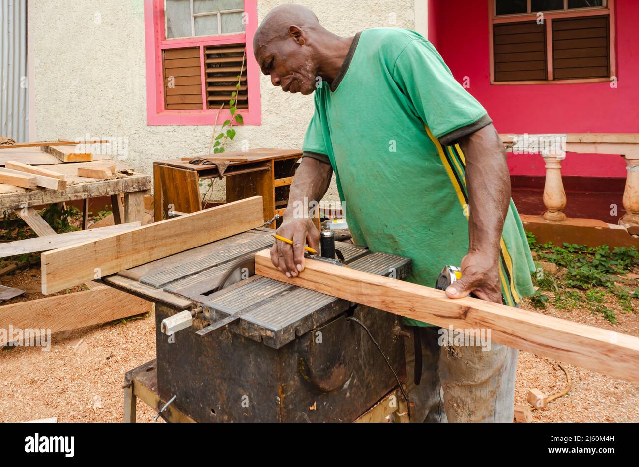 Un costruttore di cabinate con un pennel tra le sue dita sostiene un pezzo di legno 2 x 2 come usa una vecchia sega elettrica per tagliare la sua estremità Foto Stock