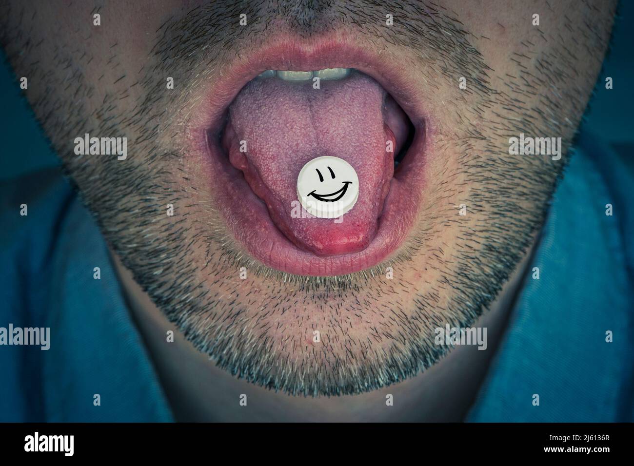 Una pillola con una droga sintetica sulla lingua di un giovane uomo. Il ragazzo succhia l'acido LSD. Faccia sorridente su una tavoletta bianca Foto Stock