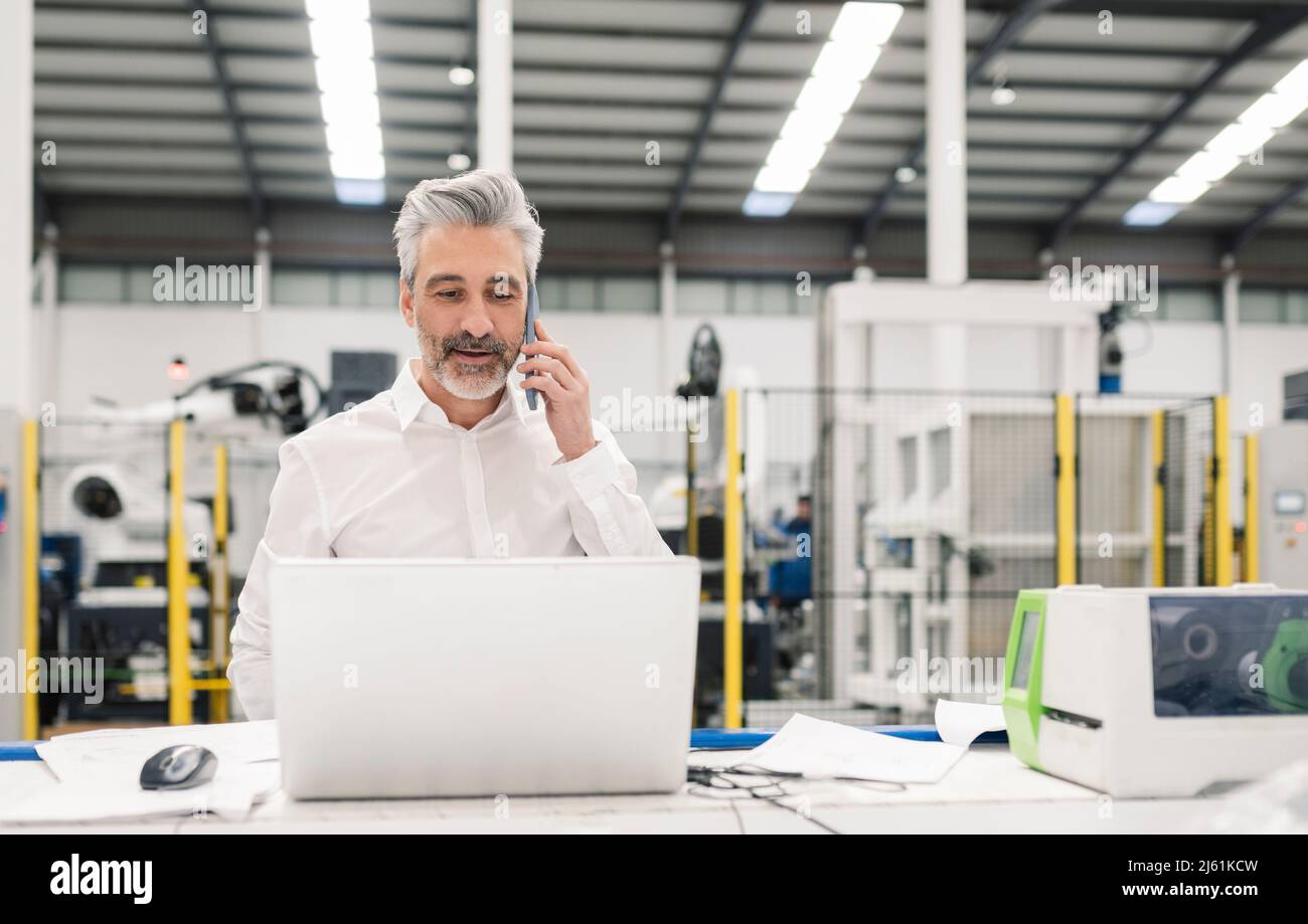 Uomo d'affari che parla su smartphone in piedi con un computer portatile alla scrivania in fabbrica Foto Stock