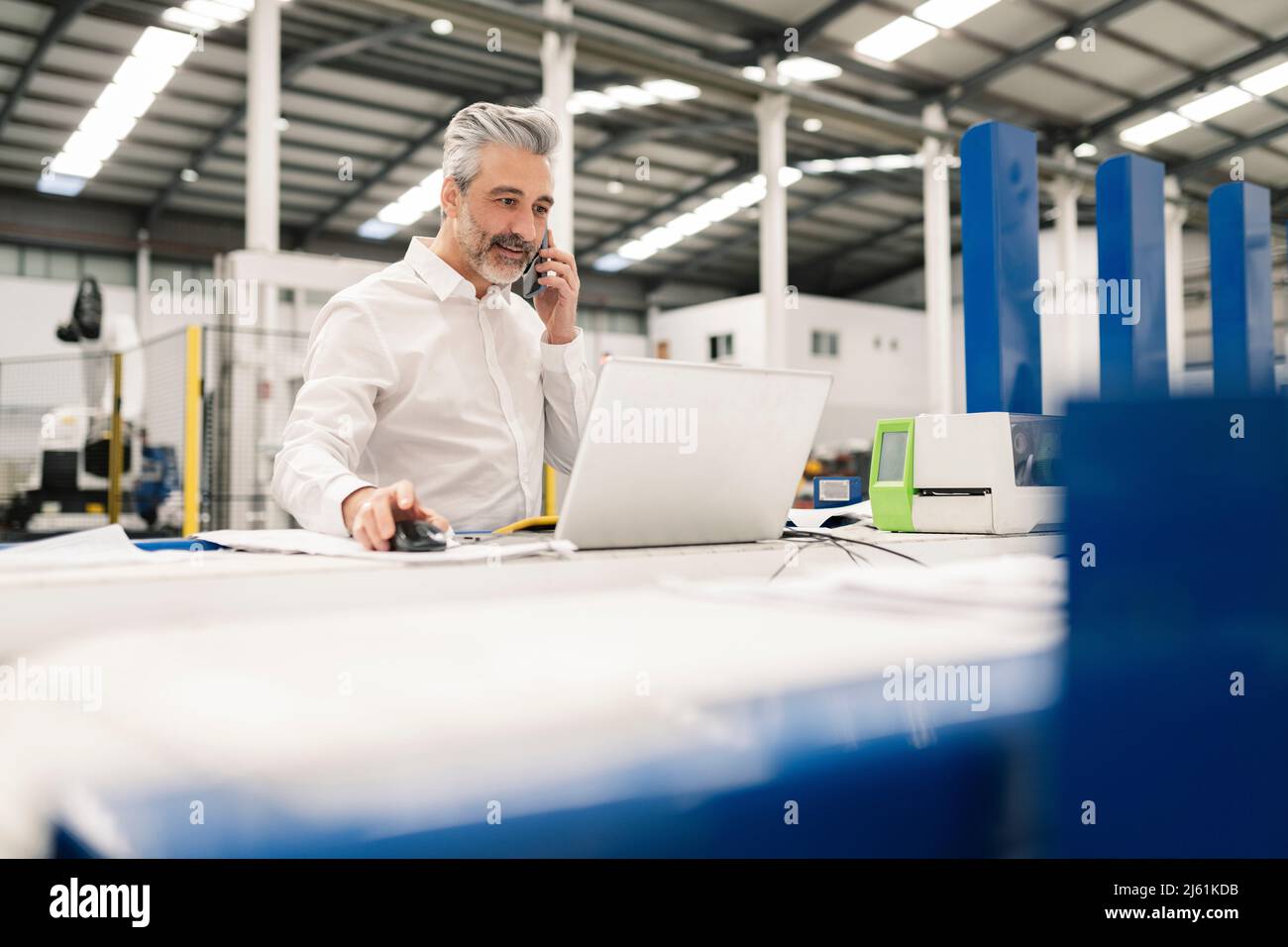 Uomo d'affari che usa il laptop che parla con lo smartphone in fabbrica Foto Stock