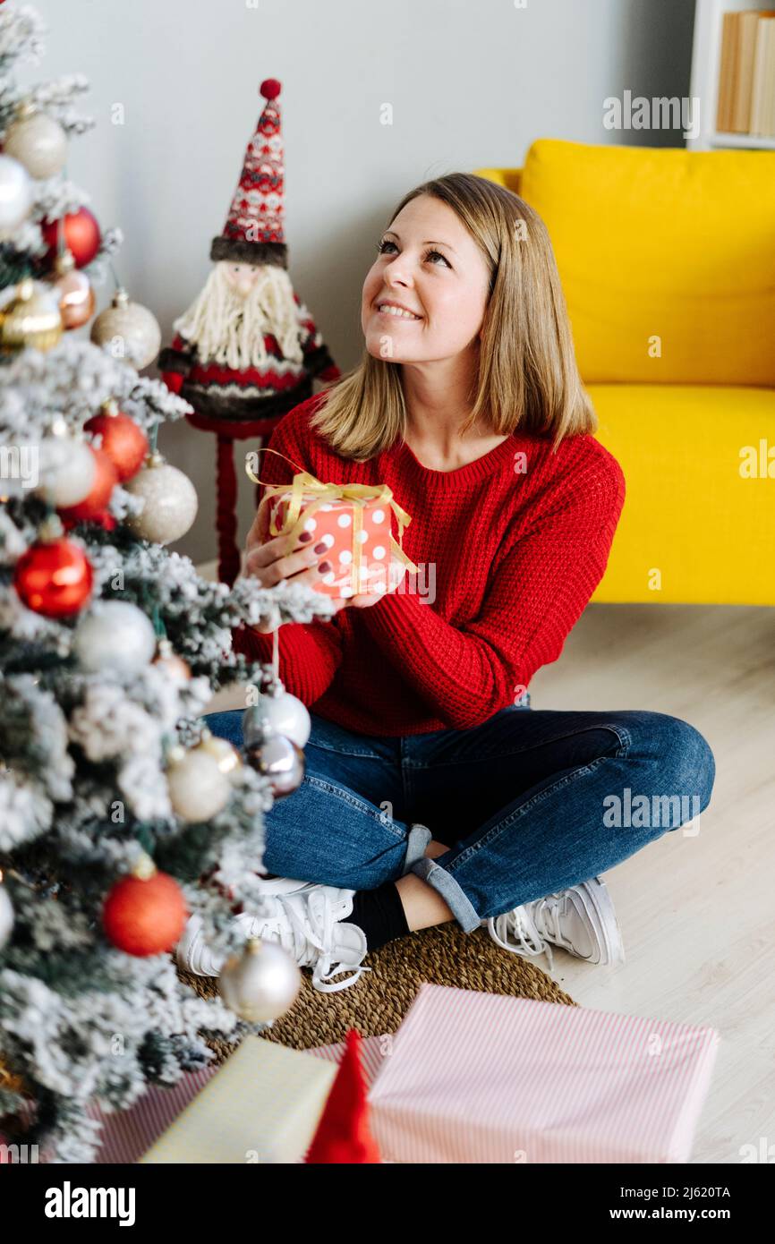 Donna sorridente con regalo di Natale guardando l'albero decorato in soggiorno a casa Foto Stock