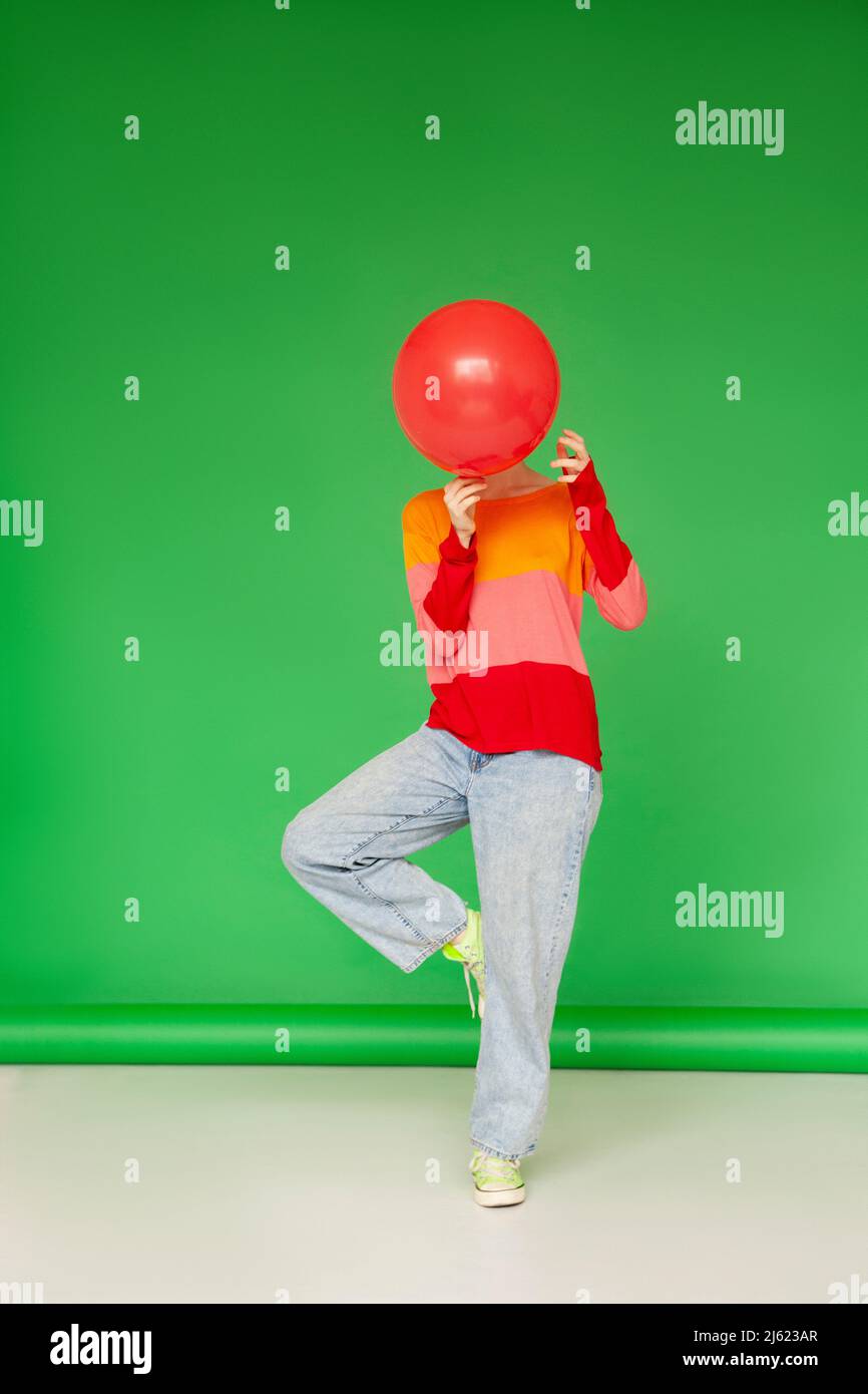 Donna che ricopre il viso con palloncino rosso in piedi su una gamba su sfondo verde Foto Stock