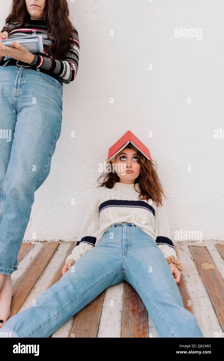 Donna annoiato con libro sulla testa reclinato di fronte al muro bianco sul cortile posteriore Foto Stock