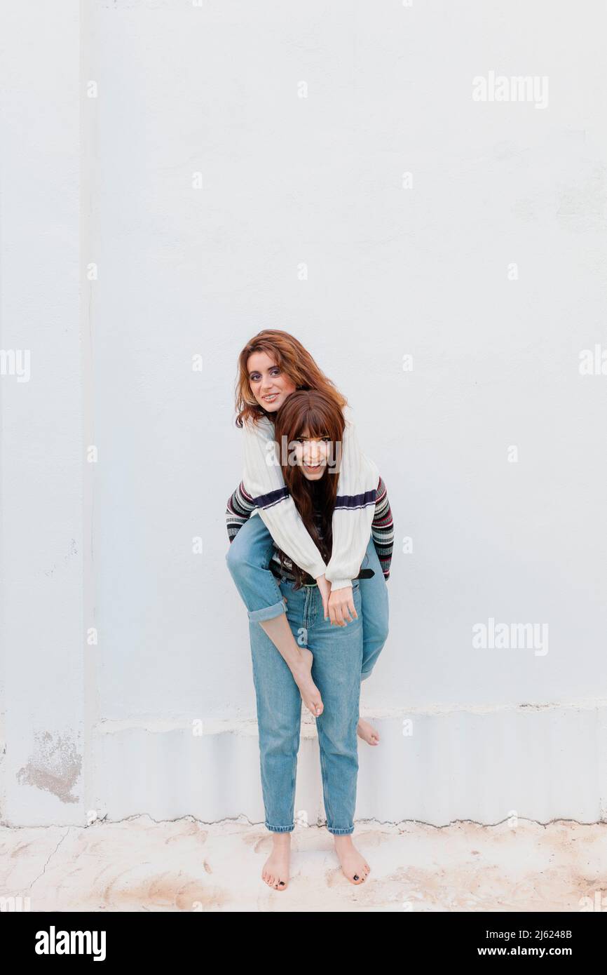 Felice donna piggybacking amico di fronte alla parete bianca a cortile posteriore Foto Stock