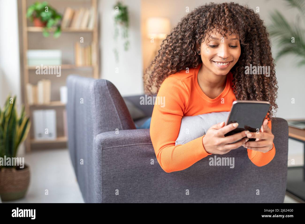 Donna sorridente che invia messaggi di testo tramite smartphone sul divano in salotto Foto Stock