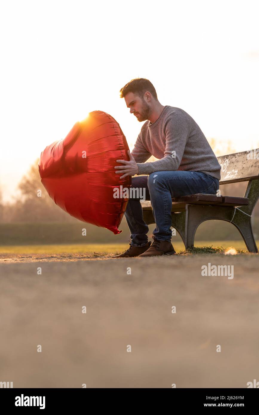 Triste giovane uomo che tiene su un pallone a forma di cuore seduto su una panchina nel parco Foto Stock