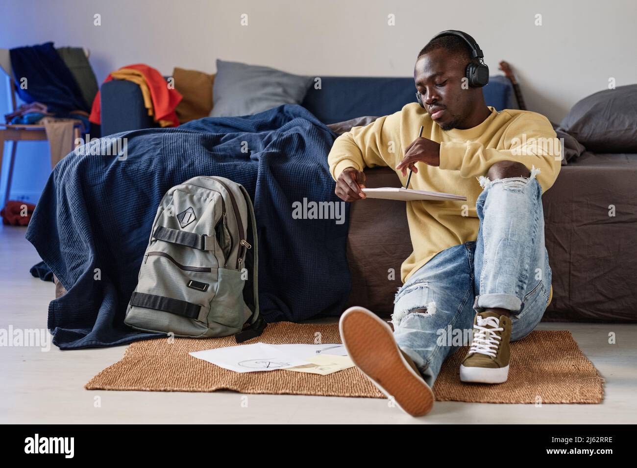 Serio rilassato giovane uomo nero in cuffie seduto sul pavimento e appoggiato sul divano mentre disegnano lo schizzo nella stanza dello studente Foto Stock