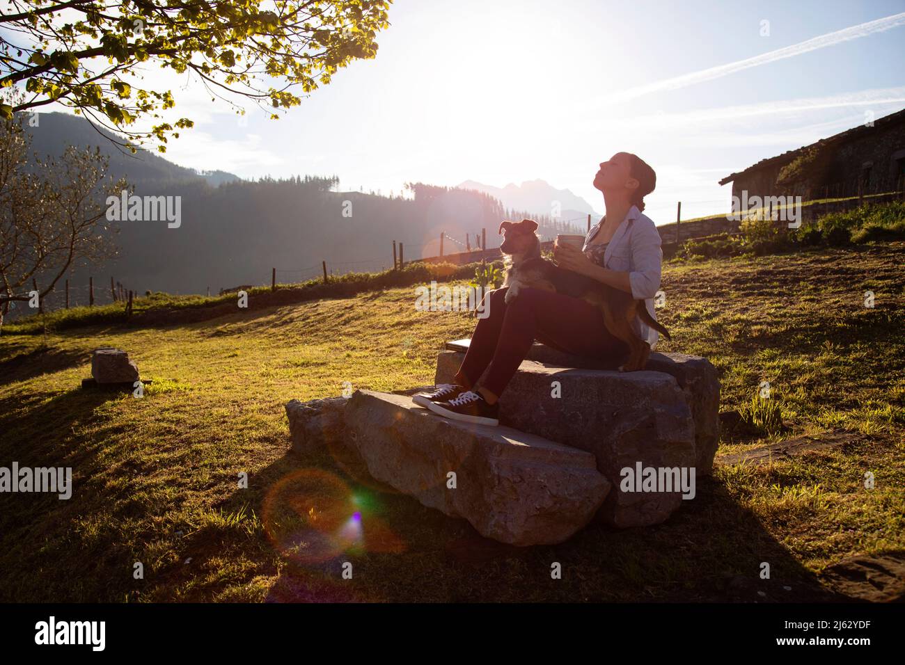 giovane donna che gode i raggi del sole al tramonto con una tazza di caffè in mano e accompagnato dal suo cane bodeguero. zona rurale. stagione delle vacanze. relax Foto Stock