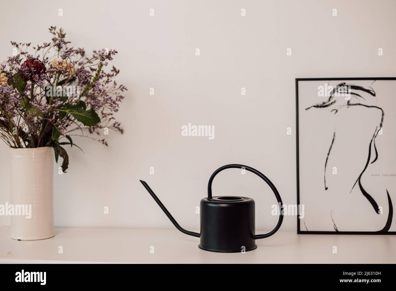 Un irrigatore a fiore nero si erge su una mensola bianca vicino a fiori e dipinti Foto Stock