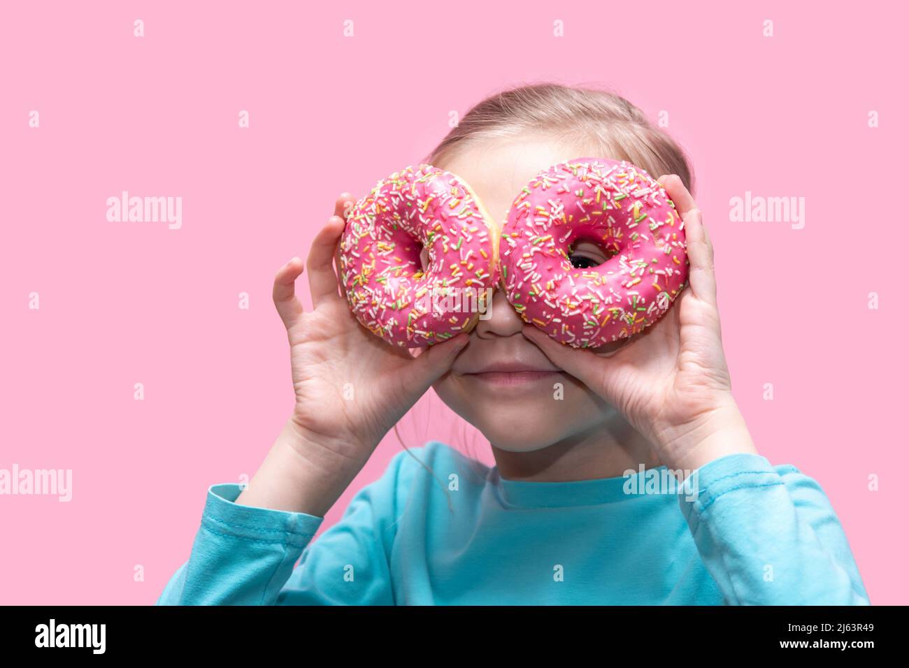 Una ragazza divertente carina in una t-shirt blu tiene le ciambelle rosa brillante vicino ai suoi occhi come gli occhiali e vi guarda su uno sfondo rosa. Conc. Infanzia allegra Foto Stock