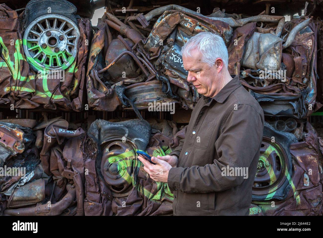 Un uomo che controlla il suo telefono cellulare mentre si trova di fronte a un muro di auto schiacciate. Ironia della sorte, i conducenti di queste auto potrebbero aver fatto lo stesso. Foto Stock