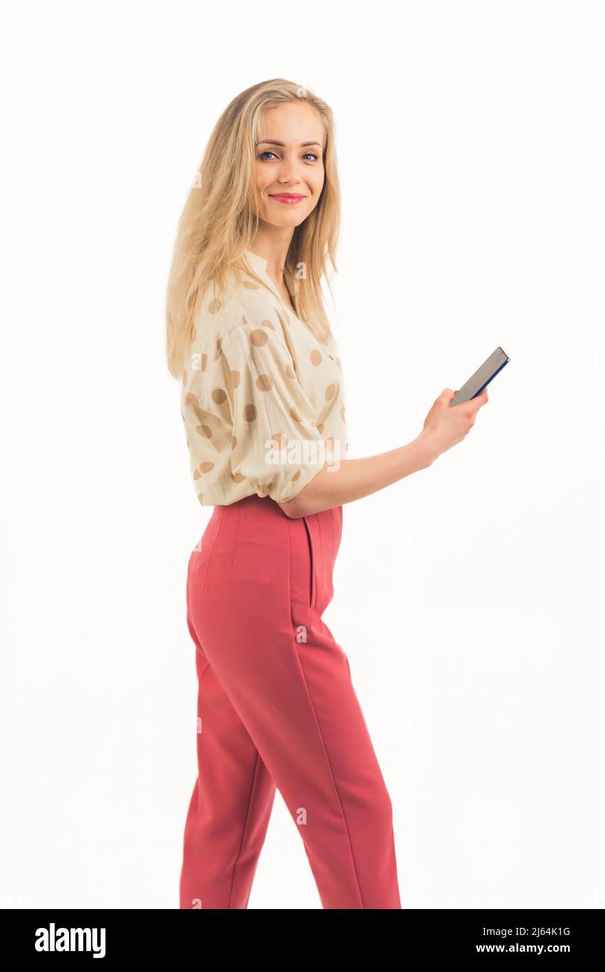 Bella ragazza bionda europea con occhi blu sorridente indossando una camicia e pantaloni rosa in piedi lateralmente e tenendo il telefono studio girato bianco sfondo medio pieno scatto copia spazio. Foto di alta qualità Foto Stock