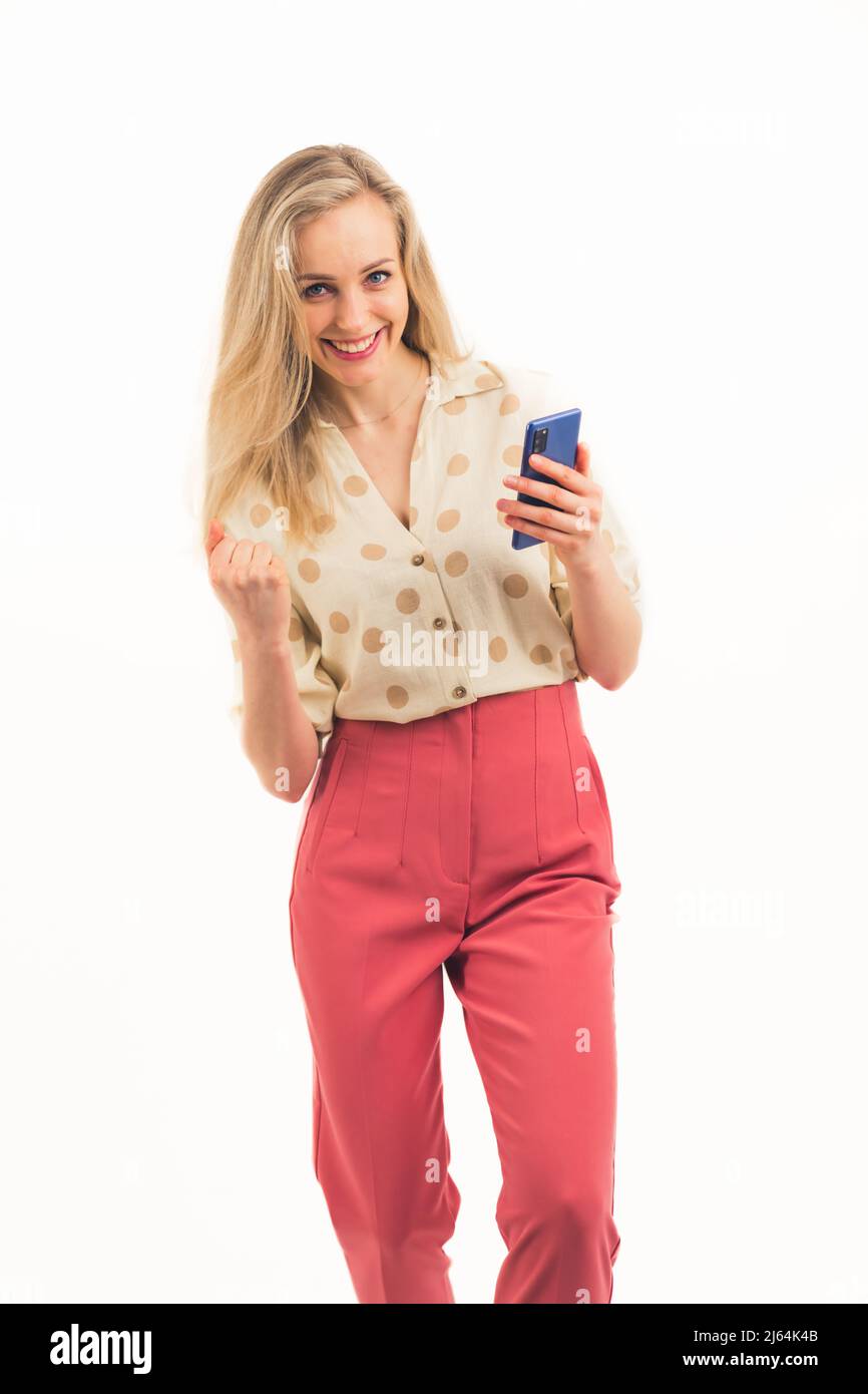 Affascinante ragazza caucasica bionda blu-eyed con un pugno di grinning clenched e tenendo un telefono che indossa una camicia e pantaloni rosa medio full shot bianco sfondo copia spazio scatto studio. Foto di alta qualità Foto Stock