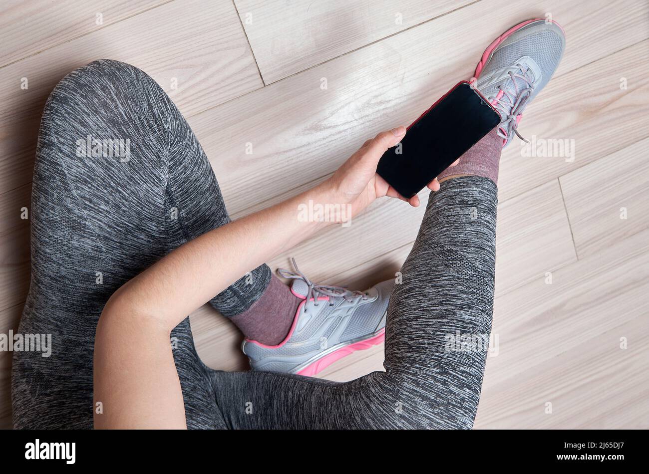 Donna in abbigliamento sportivo e scarpe sportive con telefono cellulare seduto sul pavimento, piatto. Pronto per l'allenamento in palestra Foto Stock