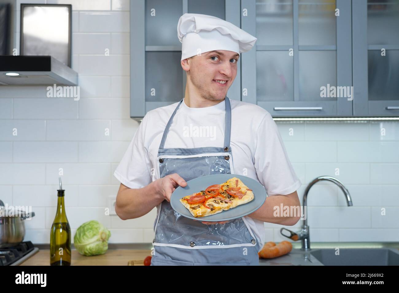 Ritratto di uno chef professionista in cucina. Tiene in mano una pizza cotta e una bottiglia di olio d'oliva. Foto Stock