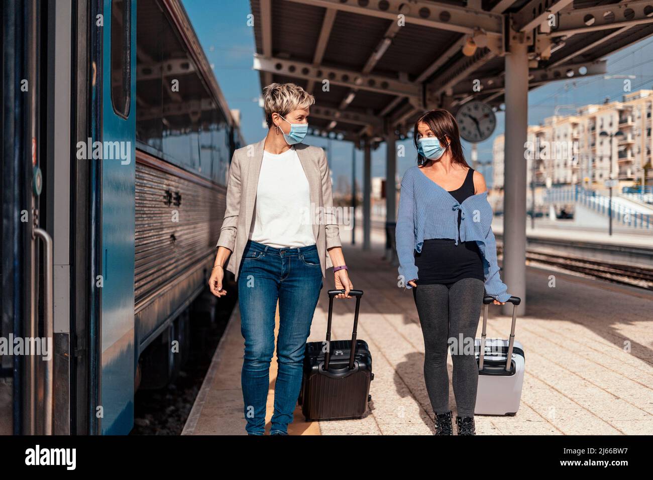 Due allegri amici femminili in maschere protettive camminando e chiacchierando sulla piattaforma alla stazione ferroviaria Foto Stock