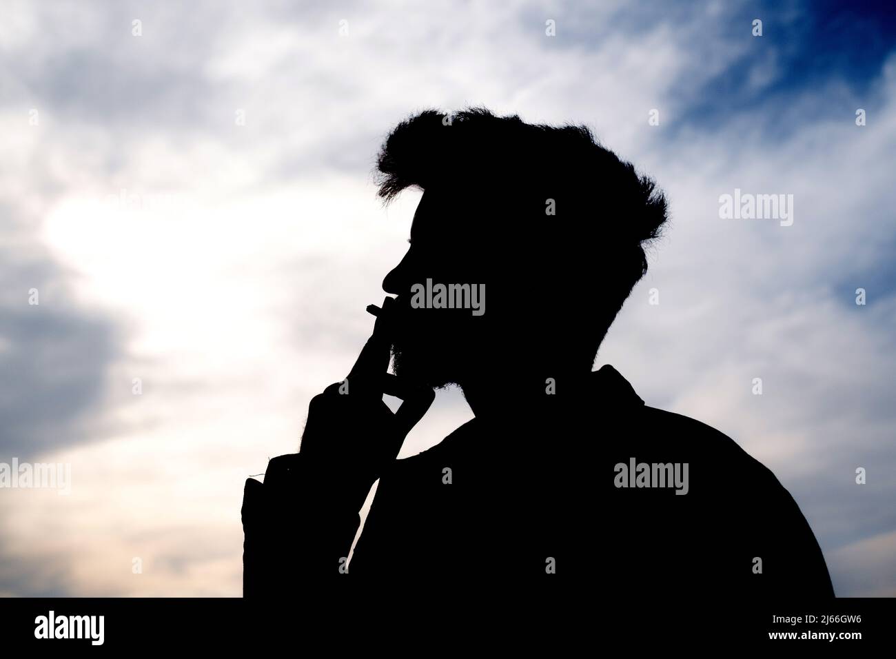 Una silhouette da uomo giovane che fuma. Un uomo caucasico ritratto fumo sigaretta silhouette su una panca parco all'aperto. Pensare alla vita, uomo triste. Foto Stock