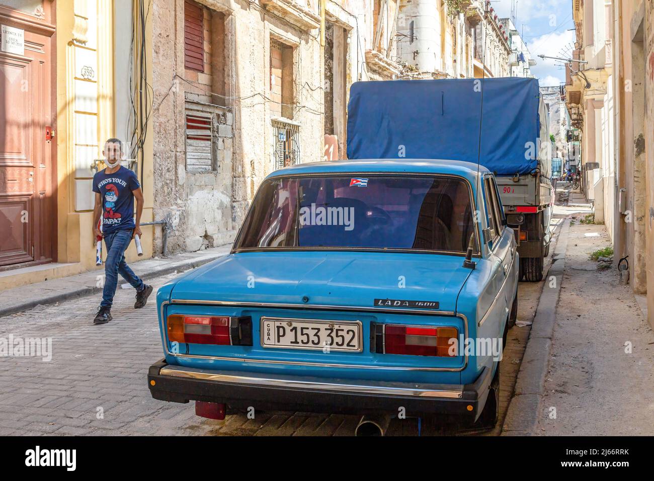 Un uomo cubano cammina in una strada acciottolata con edifici meteoriti su entrambi i lati. Nel quartiere residenziale è parcheggiata una macchina blu russa Lada. Foto Stock