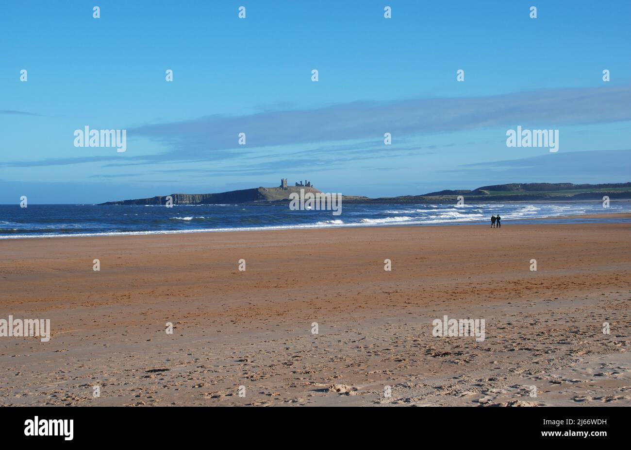 Un'ampia spiaggia aperta e quasi vuota con due persone lontane che camminano lungo la costa e il castello di Dunstanburgh sul suo crinale come sfondo distante Foto Stock