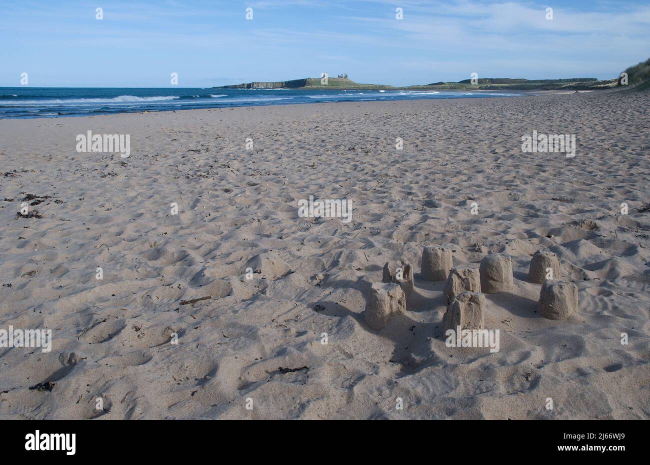 Spiaggia ampia e aperta senza gente con un piccolo castello di sabbia in primo piano e un lontano Dunstanburgh Castello sulla sua scarpa in primavera Foto Stock