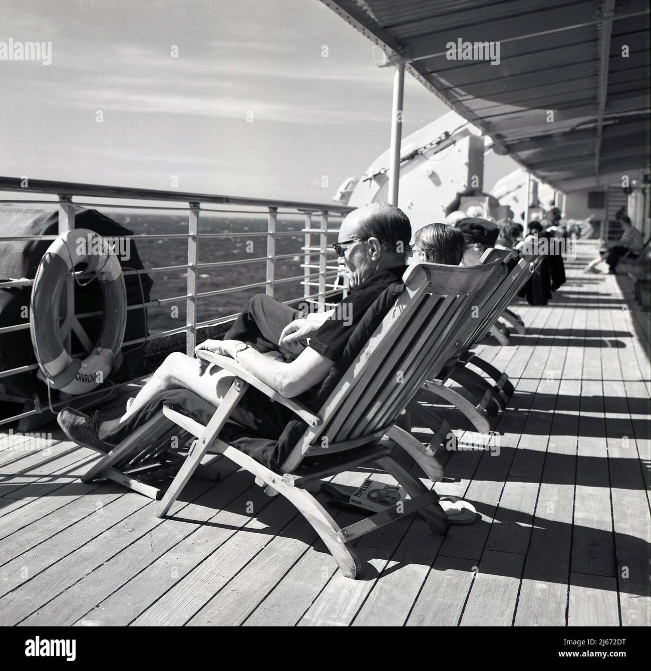 1960s, storico, medio oceano e passeggeri rilassarsi in sedie a sdraio in legno incorniciato sul ponte della nave da crociera Sagafjord guardando verso il mare. Foto Stock