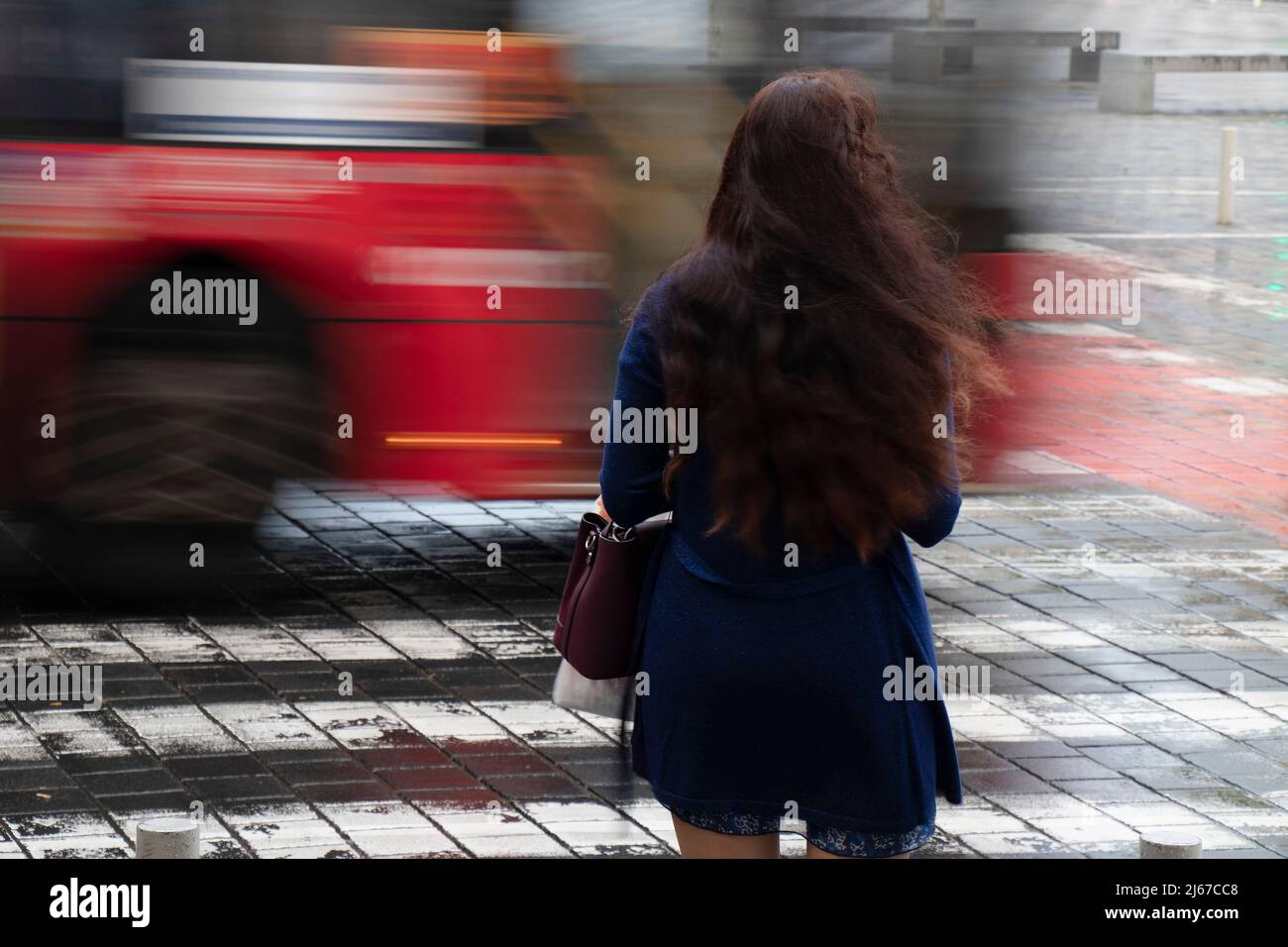 Giovane donna con lunghi capelli ondulati in attesa su un incrocio in una giornata piovosa con traffico in movimento sfocato, vista posteriore Foto Stock
