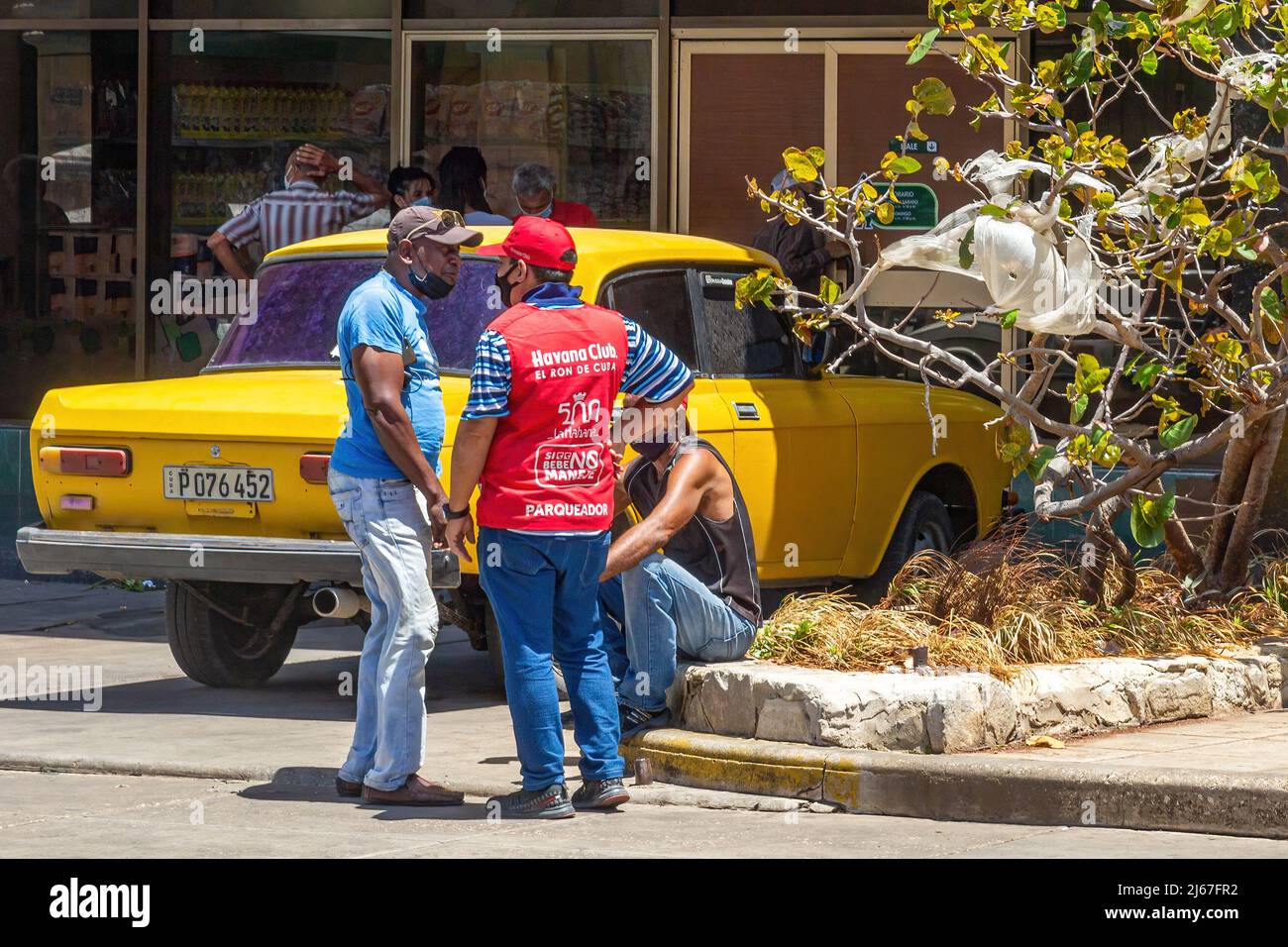 Un parcheggiatore (giubbotto rosso) parla con altri due uomini cubani che indossano maschere facciali. Un'auto gialla Moskvitch è parcheggiata in un negozio MLC Foto Stock