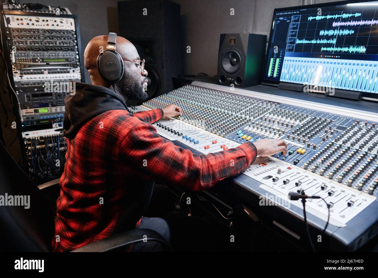 Elegante produttore di musica afro-americana che indossa cuffie sedute alla scrivania di mixaggio della canzone di registrazione in studio Foto Stock
