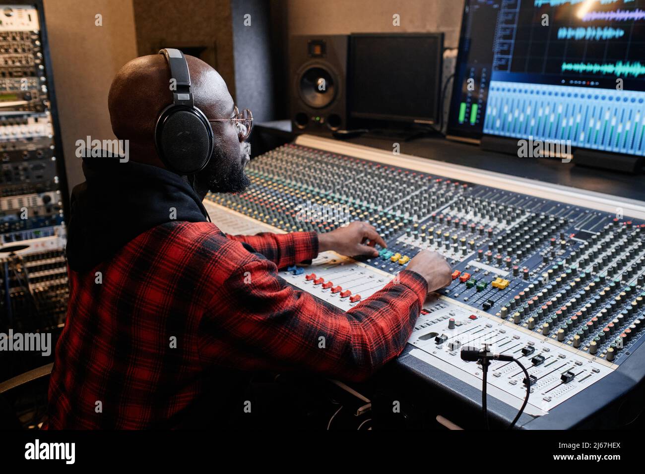 L'elegante uomo afro-americano indossa una camicia rossa e cuffie che lavorano su una traccia musicale in studio di registrazione Foto Stock
