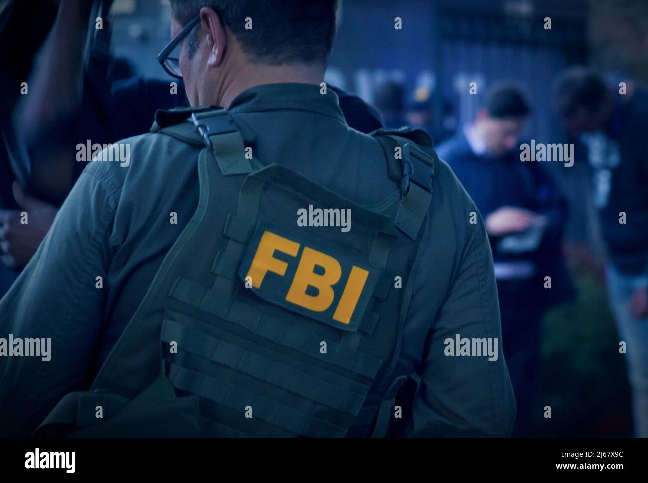 Agente FBI maschio caucasico che indossa occhiali in un gilet verde a prova di proiettile con logo FBI visto da dietro sul punto di indagine. Foto Stock