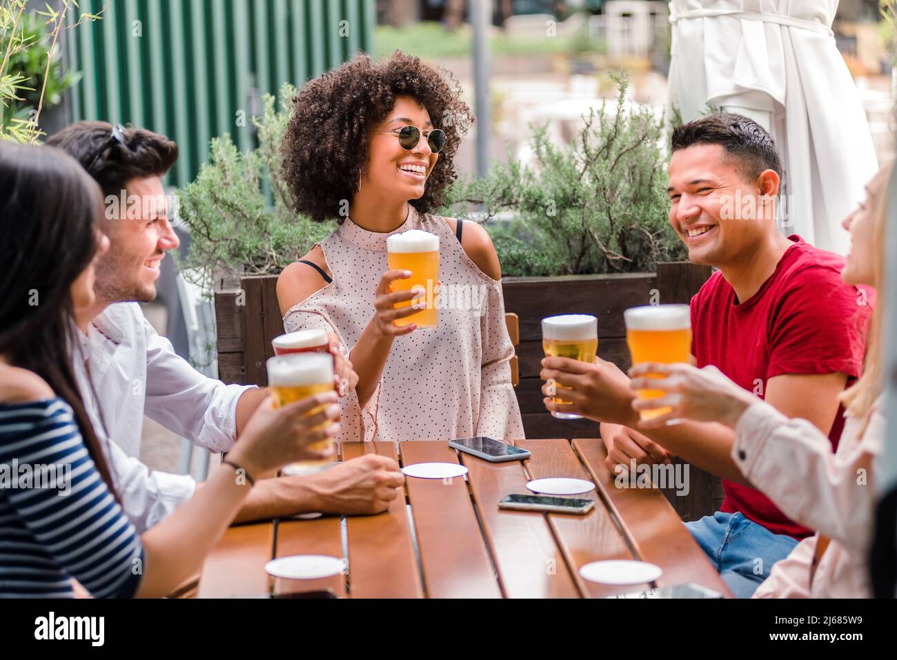 Gruppo multietnico di amici o studenti seduti a un tavolo di legno mentre tostate e bevete birra insieme all'aperto in un pub Foto Stock