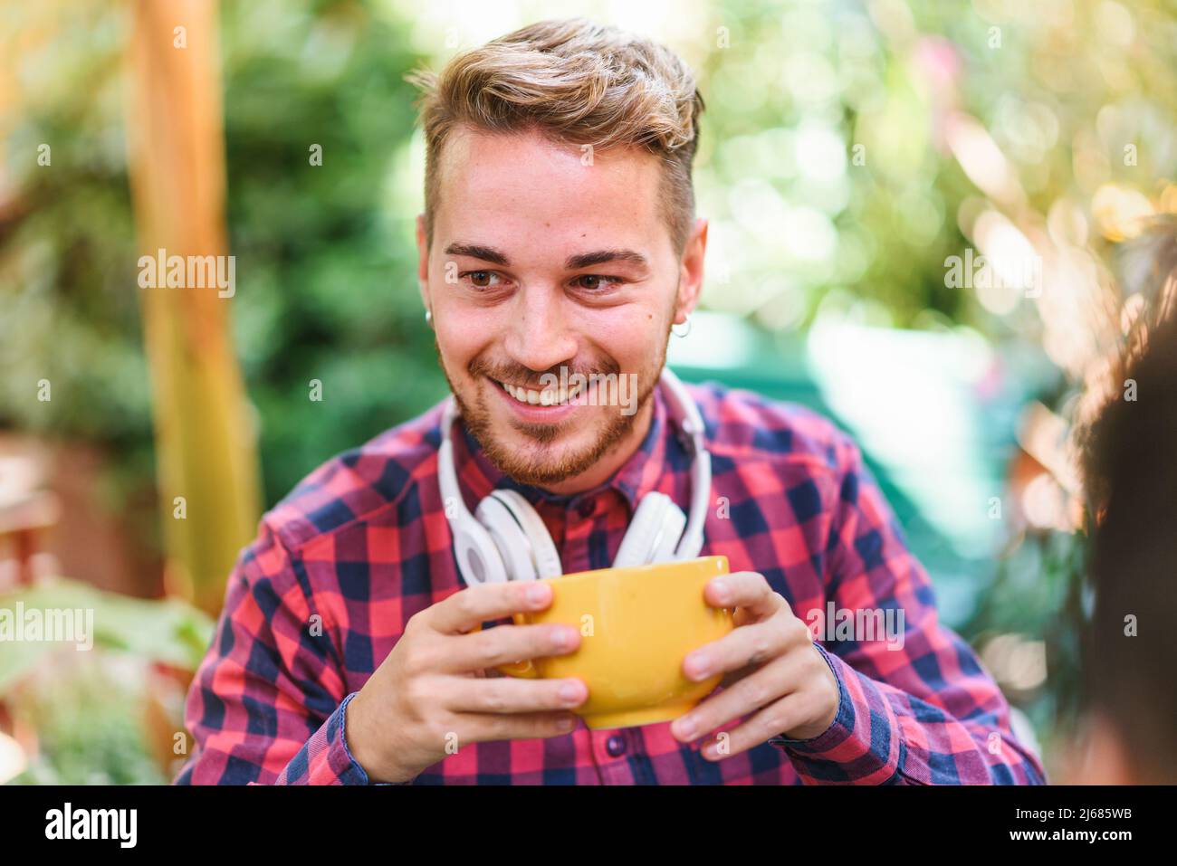 Happy maschio in camicia a scacchi sorridente e guardando via mentre si gusta il caffè fresco al mattino in cortile Foto Stock