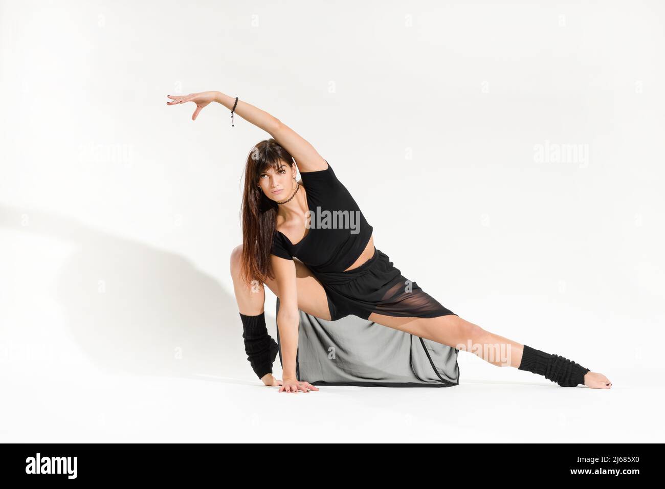 Corpo completo di giovane ballerina femminile in abito nero che si snodano davanti con instep flessibile e braccio sollevato su sfondo bianco Foto Stock
