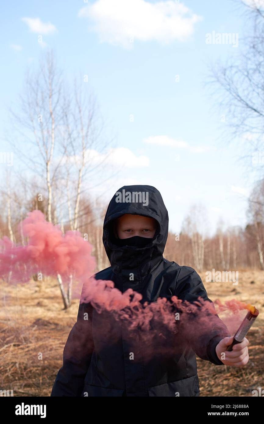 Un preteen in una giacca nera e una maschera tiene una scacchiera con fumo colorato Foto Stock