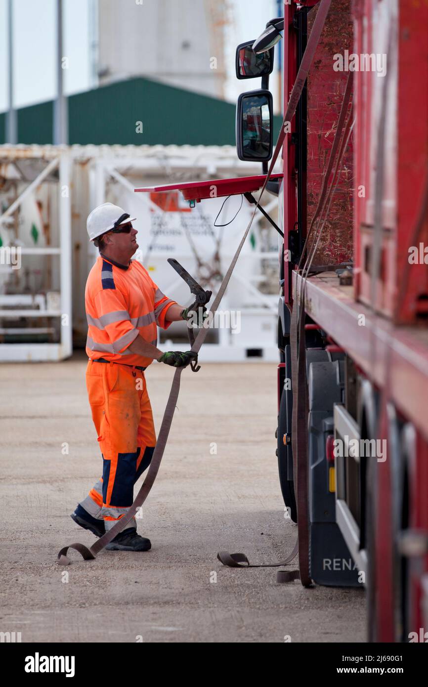 Conducente di autocarri che utilizza cinghie a cricchetto su un autocarro a pianale che trasporta container offshore Foto Stock