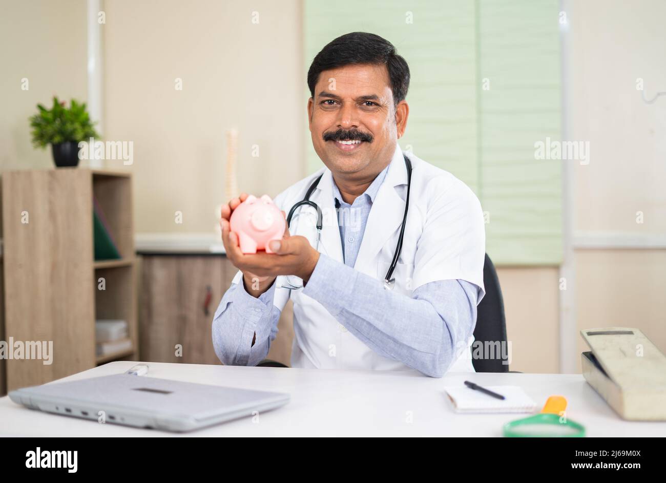 felice medico sorridente mostrando piggy banca in ospedale guardando la macchina fotografica - conept di healhcare, risparmi e assicurazione medica Foto Stock