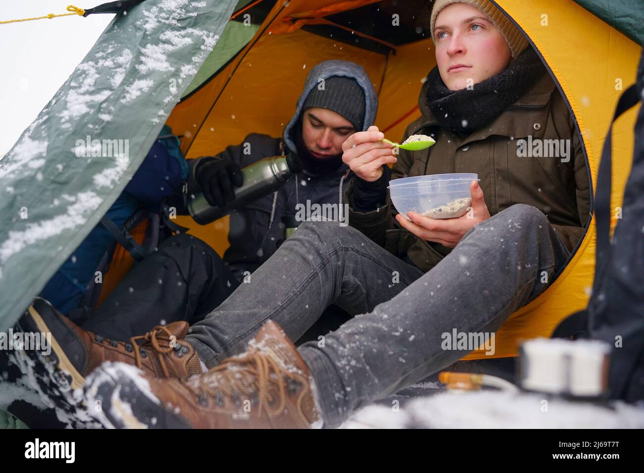 I ragazzi sono seduti in una tenda, preparando il cibo durante un'escursione invernale. Vicino alla tenda nella neve si trova la loro attrezzatura. Due uomini stanno mangiando nella tenda. C Foto Stock