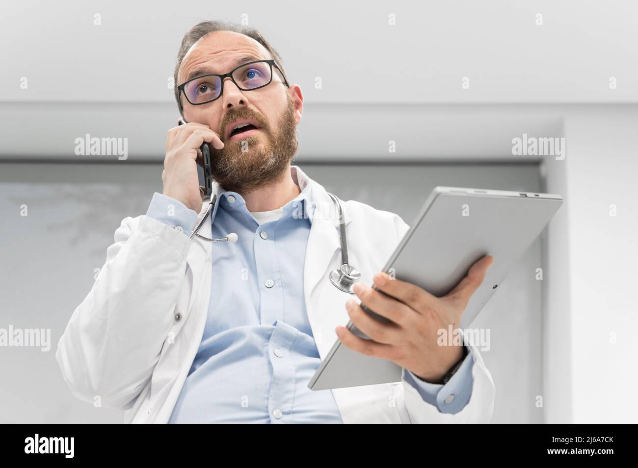 Medico di sesso maschile con stetoscopio seduto alla scrivania, parlare sul telefono cellulare e guardare il tablet digitale, discutere la diagnosi di un paziente. Fotografia di alta qualità Foto Stock