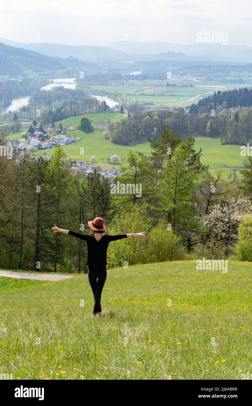 Giovane donna skinny con braccia aperte e cappello si erge su una collina nella natura della Svizzera in primavera. Foto Stock