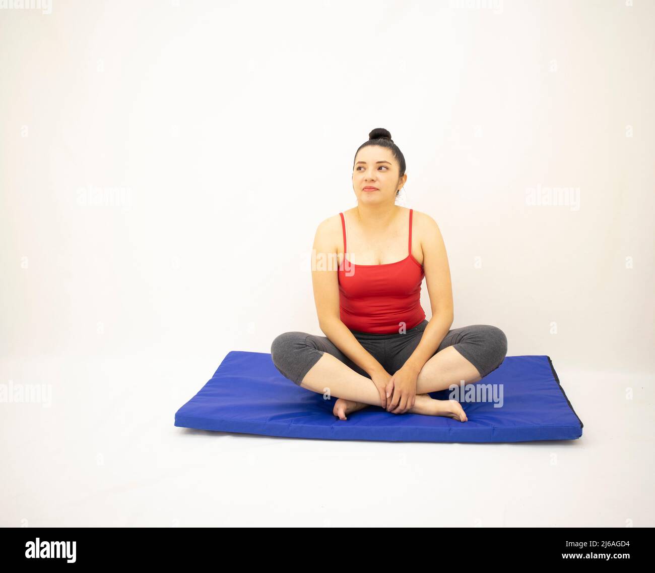 bella donna in abbigliamento sportivo grigio, blusa rossa, guarda su gambe incrociate poggianti su un tappetino blu, su sfondo bianco Foto Stock