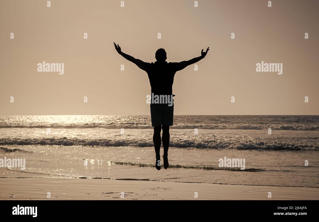 silhouette di uomo atletico che salta con mani rialzate sulla spiaggia estiva, libertà Foto Stock