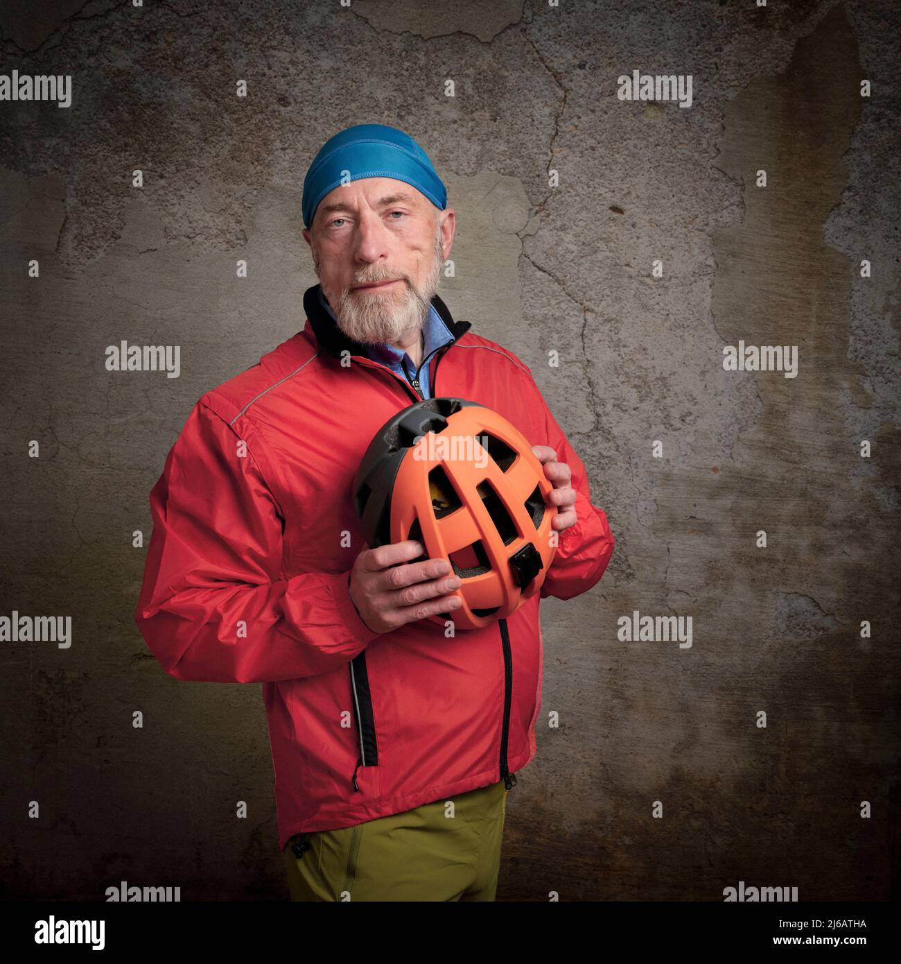 ritratto di ciclista maschile sorridente in giacca da ciclismo rosso brillante e cappello del cranio che tiene un casco contro una parete di cemento grunge Foto Stock