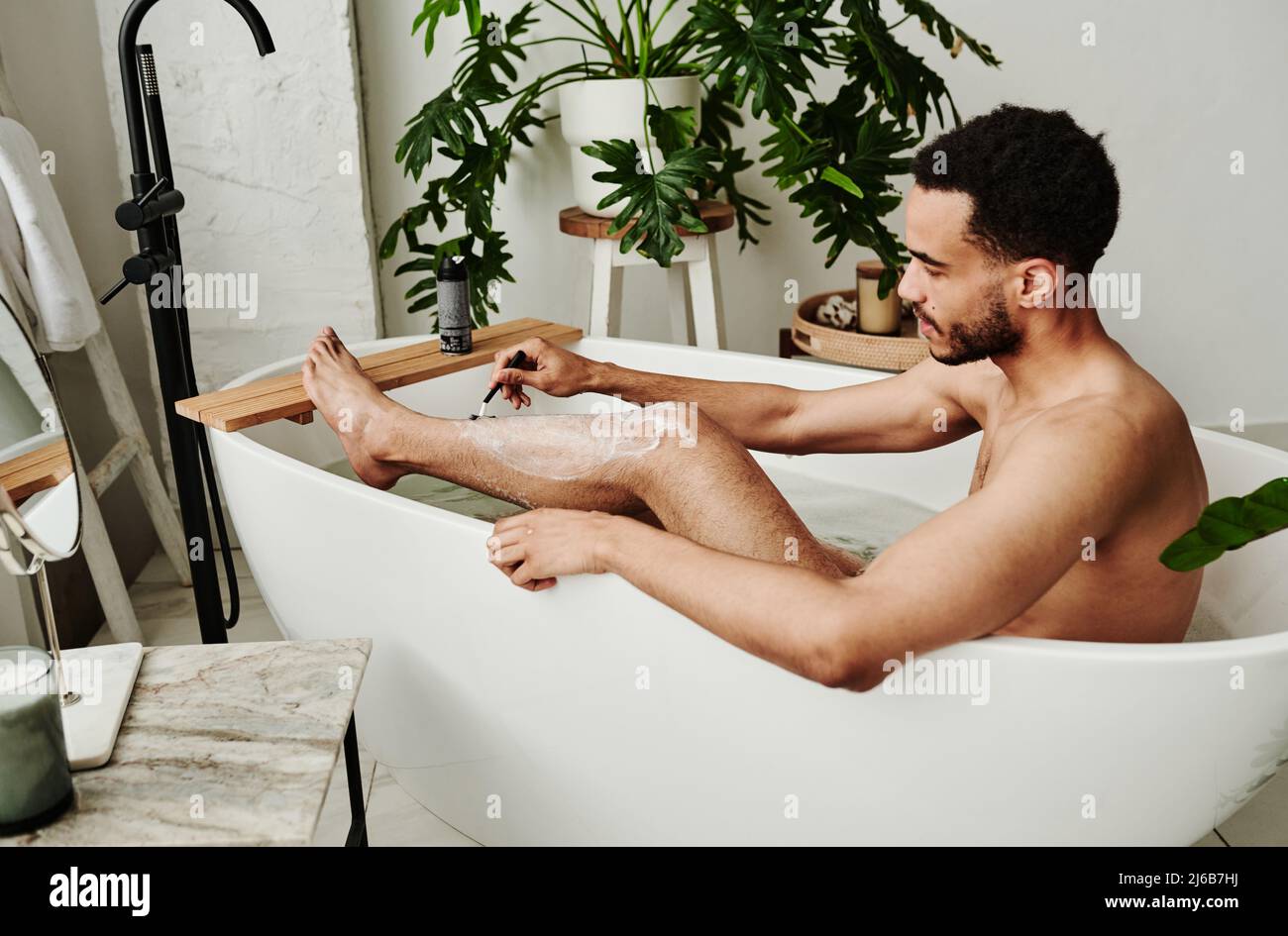 Giovane uomo omosessuale seduto in bagno e rasando le gambe durante la procedura spa Foto Stock