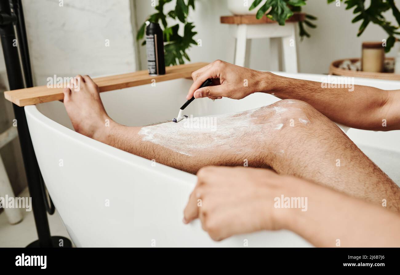 Primo piano del giovane uomo che usa il rasoio per radersi le gambe mentre  si siede in bagno Foto stock - Alamy