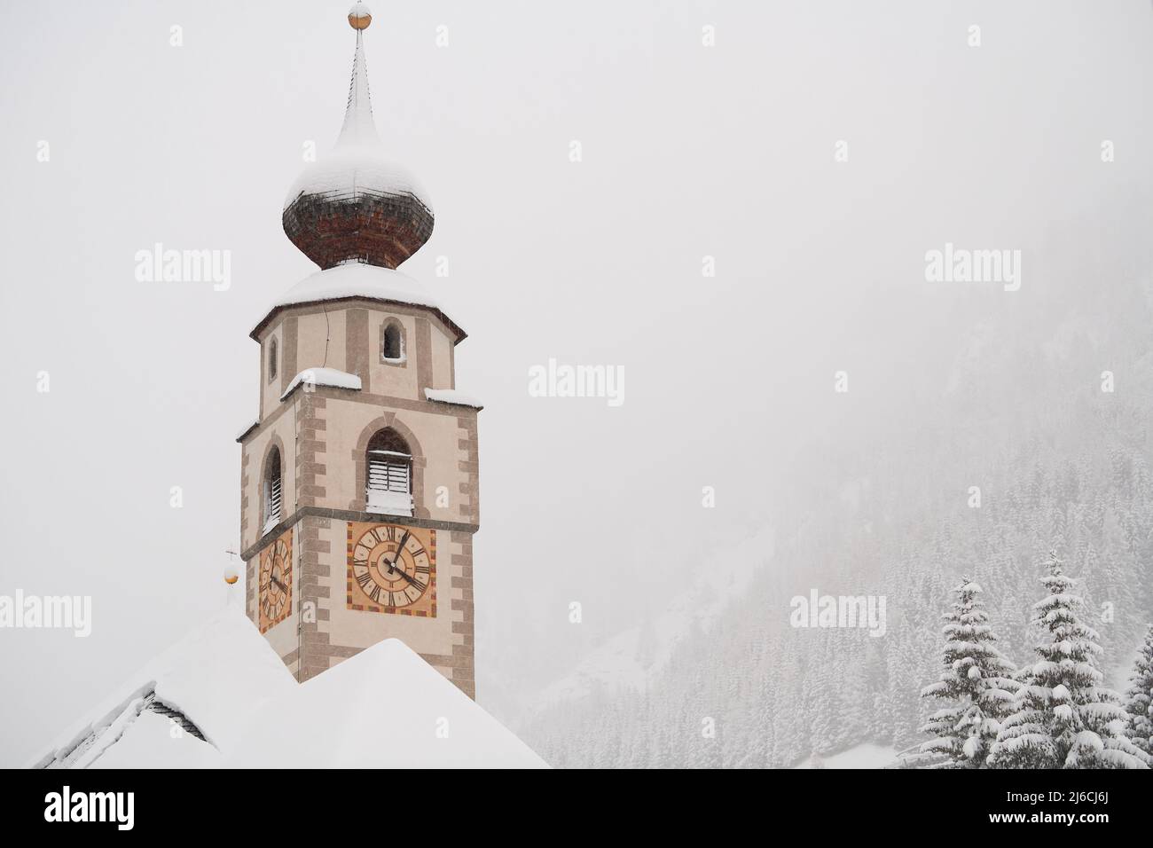 Colfosco, Italia - 5 gennaio 2022: Torre della chiesa di S. Vigil e alcuni alberi con foresta di montagna sullo sfondo con molta neve fresca. Foto Stock