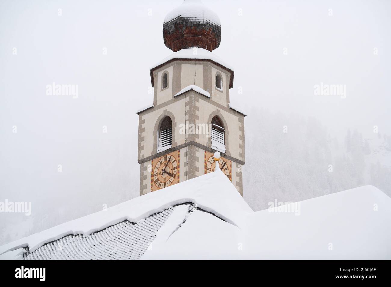 Colfosco, Italia - 5 gennaio 2022: Torre e tetto della chiesa di San Vigilio con foresta di montagna sullo sfondo con molta neve fresca. Foto Stock