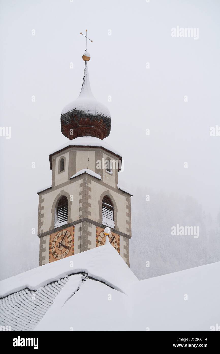 Colfosco, Italia - 5 gennaio 2022: Torre e tetto della chiesa di San Veglia con foresta di montagna sullo sfondo con molta neve fresca (verticale). Foto Stock