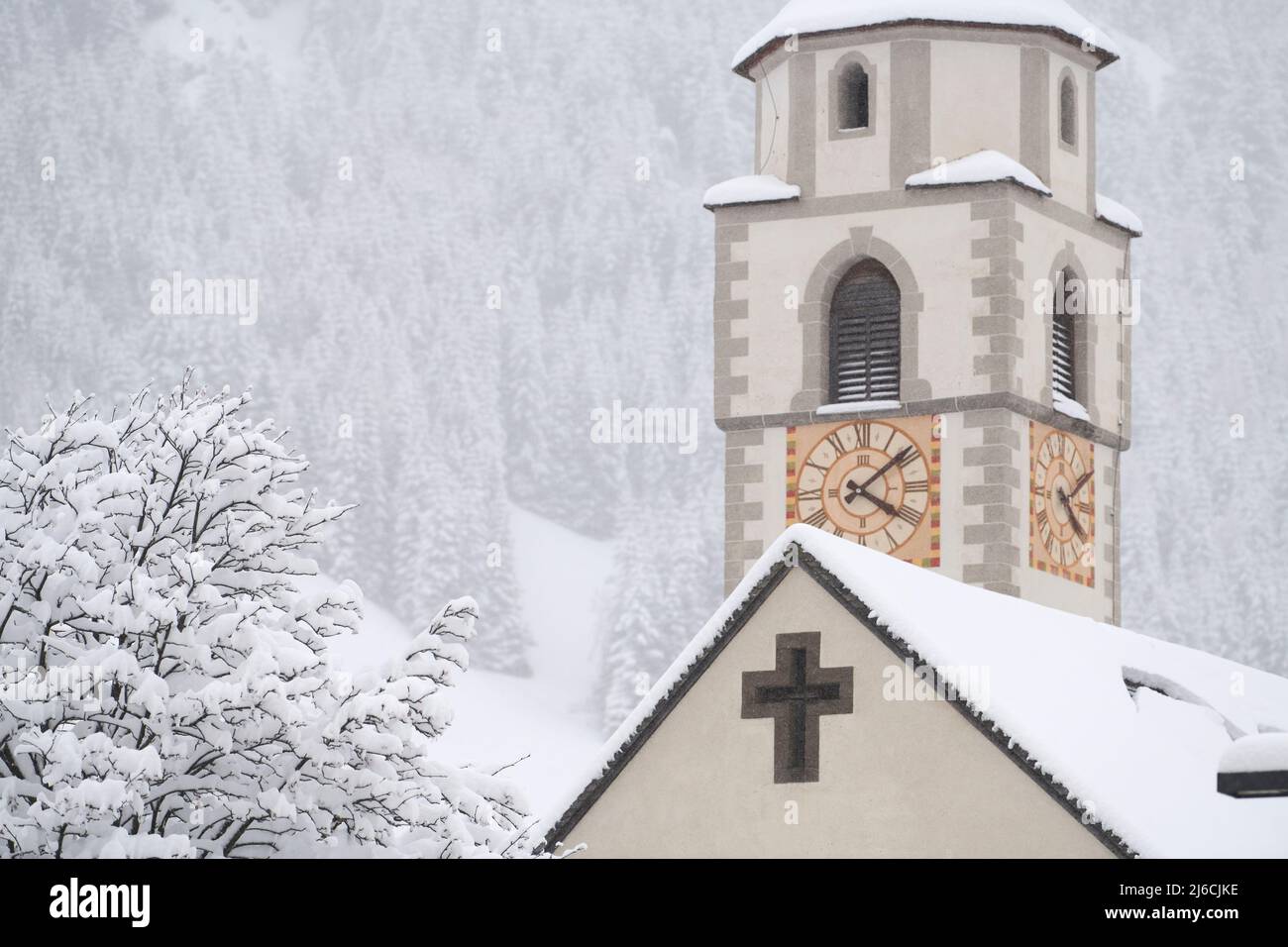 Colfosco, Italia - 5 gennaio 2022: Orologio della chiesa di S. Vigil e un unico albero con foresta di montagna sullo sfondo con molta neve fresca. Foto Stock