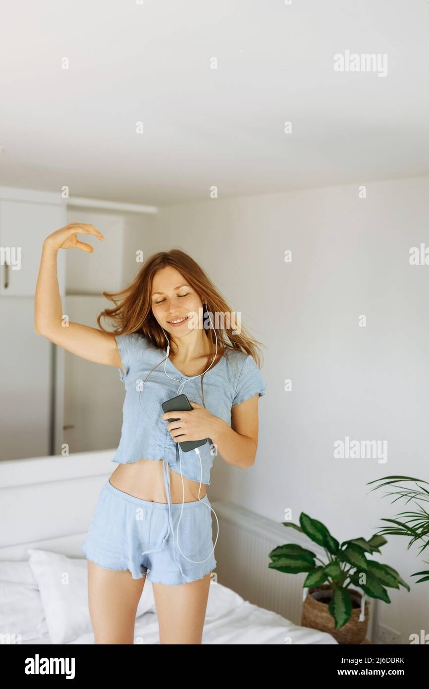 Giovane ragazza teen in pajama in piedi sul suo letto e cantare una canzone. Ragazza allegra caucasica adolescente con cuffie che ballano nella sua camera da letto . Foto Stock