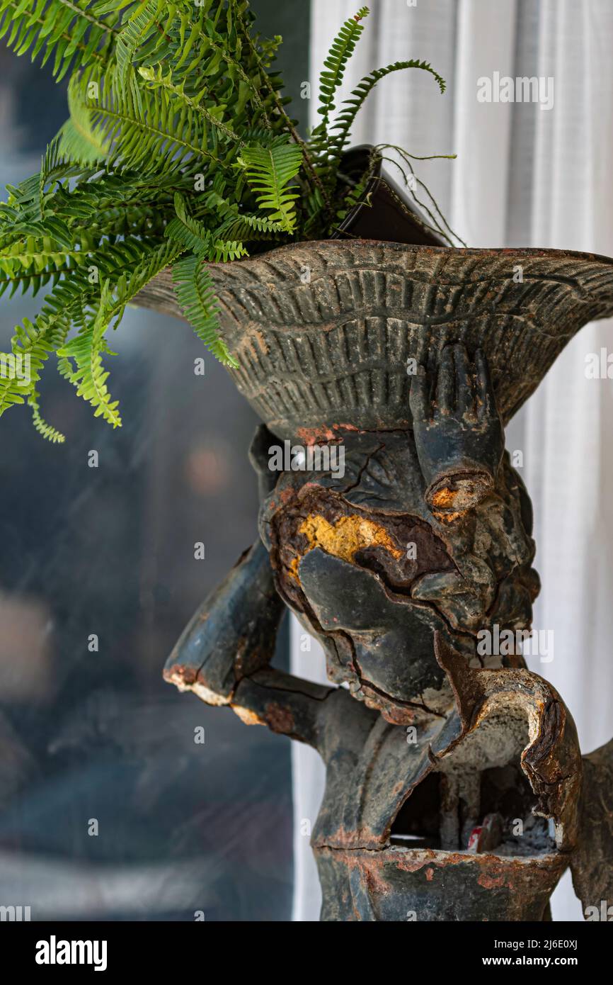 Sfondo di un contenitore di piante di cherubino rotto e arrugginito con un felce in vaso inclinato sopra con bokeh. Foto Stock